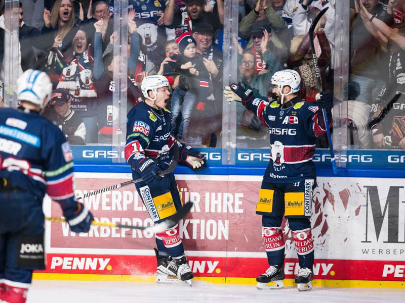 Drei Spieler der Eisbären jubeln auf dem Eis in voller Sportausrüstung. Im Hintergrund sieht man das Publikum hinter der Scheibe.