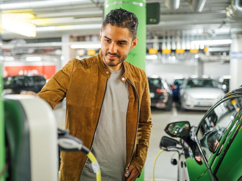 Ein junger Mann steht in einem Parkhaus an einer E-Ladestation. Neben ihm steht sein Auto und wird geladen.