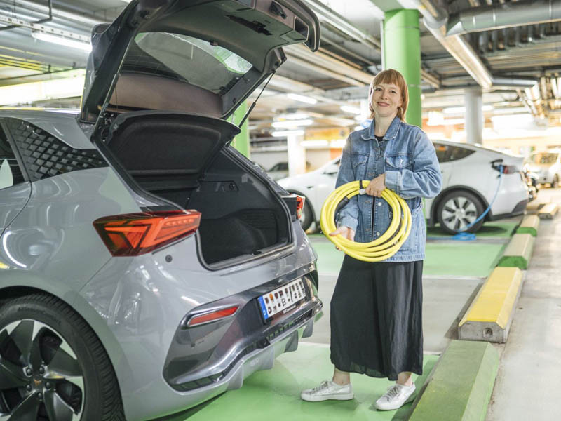 Eine junge Frau steht in einem Parkhaus am offenen Kofferraum ihres Autos und hält ein Ladekabel für das E-Laden in der Hand.